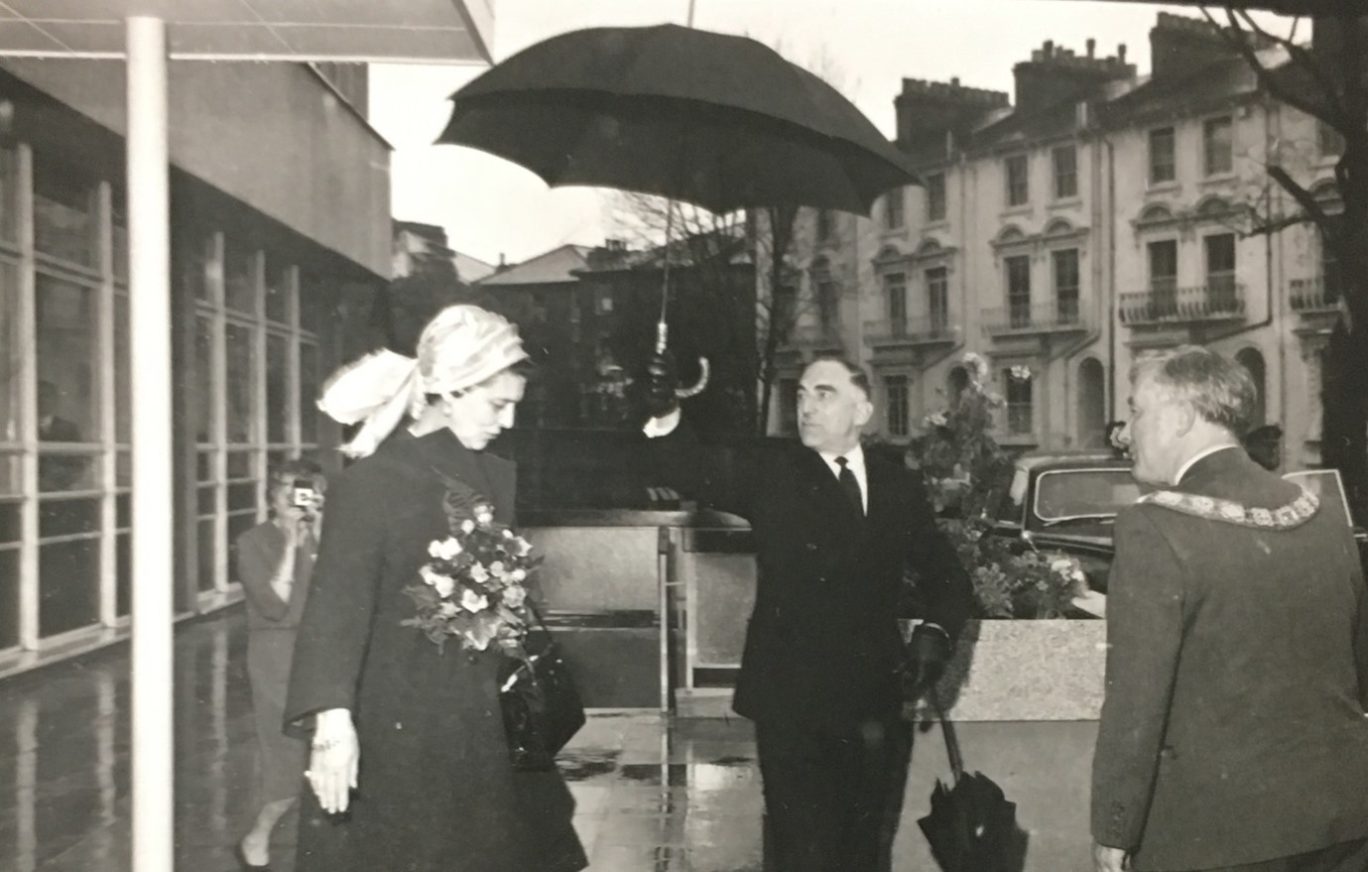Black and white image of a lady in a scarf with a man holding an umbrella over her head outside the Tavistock Centre