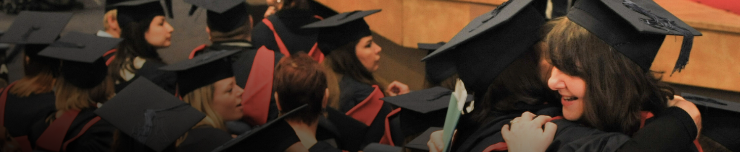 Two students in gowns and mortar board hats embrace at graduation ceremony with other graduates seated around them