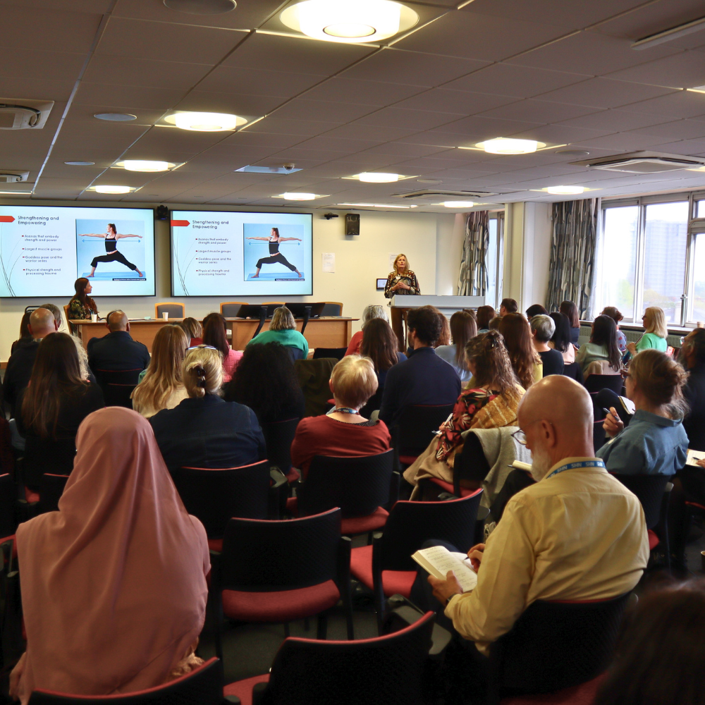 View of the lecture theatre at the Tavistock centre. The audience are listening to a presentation. The speaker is at the front and there is a presentation slide on yoga.