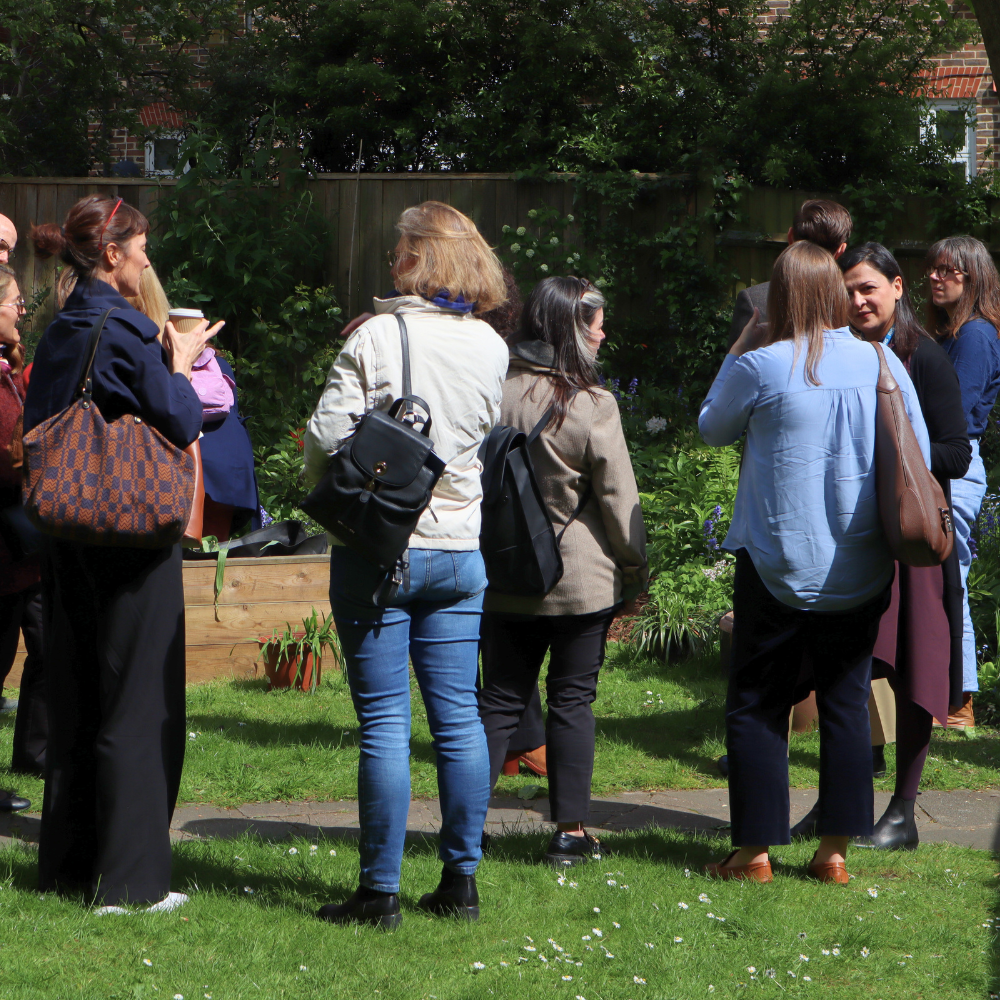 Group conversation in the garden at the Tavistock Centre. Delegates are stood up, chatting on the grass and there are flower beds behind them.