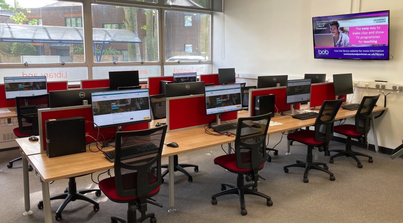 A room of computers with multiple desks, each equipped with a monitor, keyboard, and mouse. The chairs are black and red office chairs. On the right-hand wall, a large screen displays information about a library service. The room is well-lit with natural light coming from large windows on the left side.