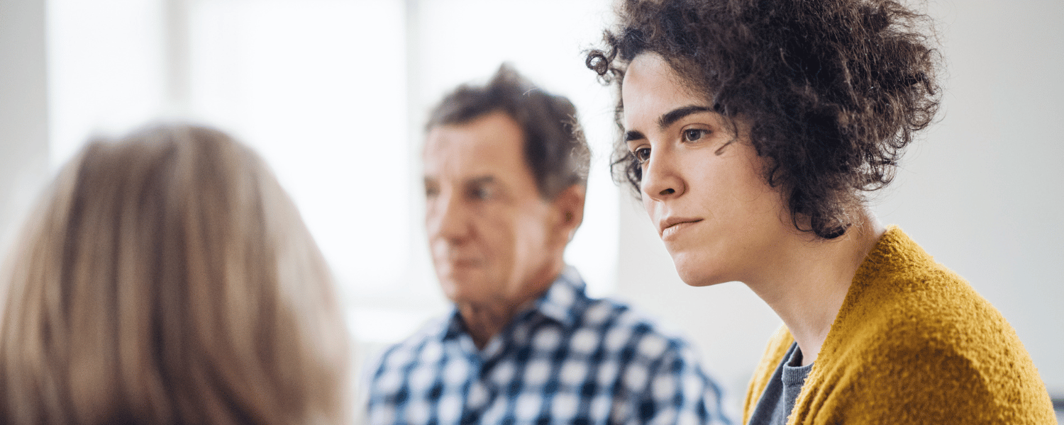 Serious woman in yellow cardigan listens to a colleague.