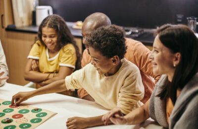 A smiling boy places a chip on a board game whilst playing it with his family. There are two adults and a young girl smiling and sat around the table with him.