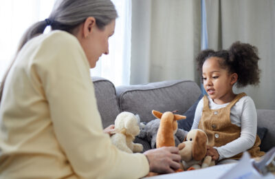 A woman and a young child sit opposite each other on a grey sofa playing with soft toys.