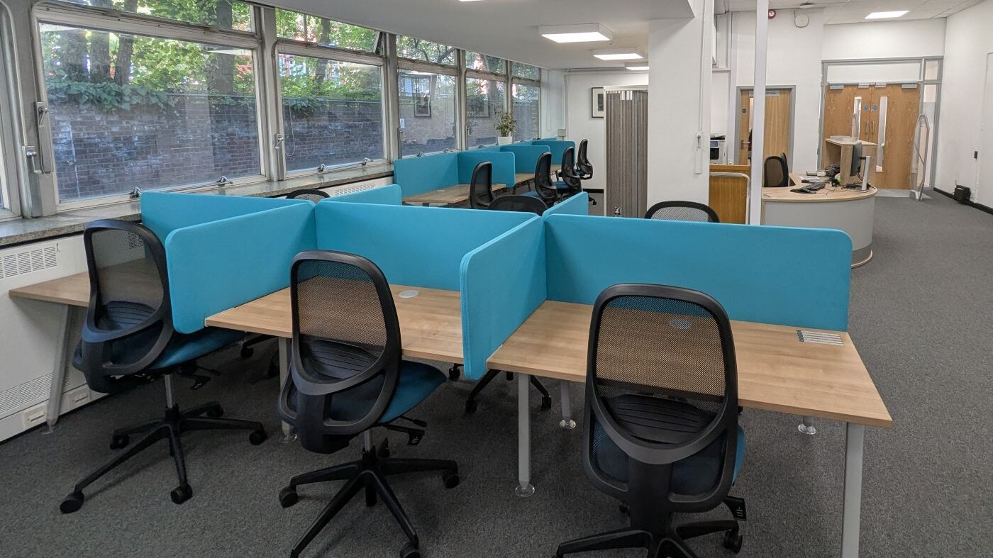 Library main space with a row of study desks with chairs in the foreground, and a row behind the library reception desk to the left